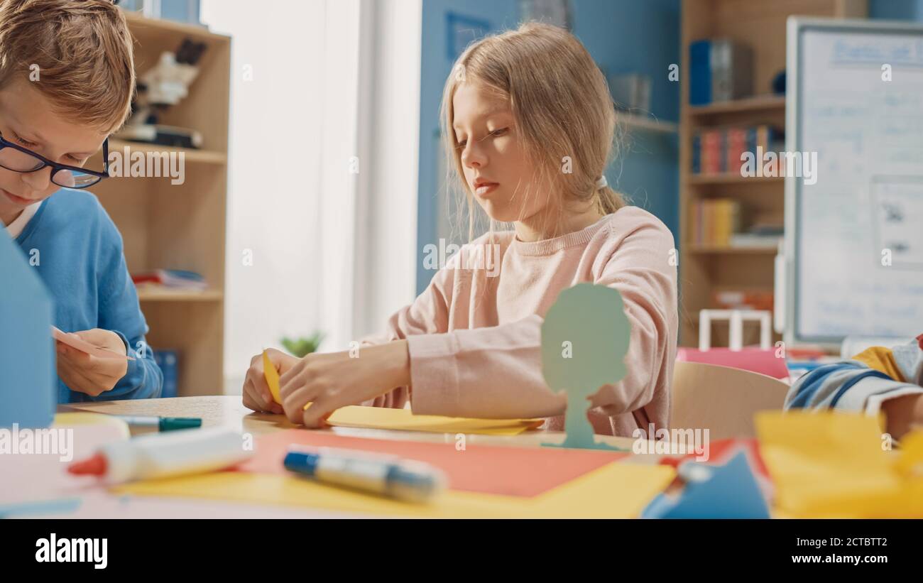 Grundschule Kunst Handwerk Klasse: Niedliche kleine Blonde Mädchen hat Spaß mit ihren Klassenkameraden auf dem Handwerk-Projekt, verwendet buntes Papier zu falten Stockfoto