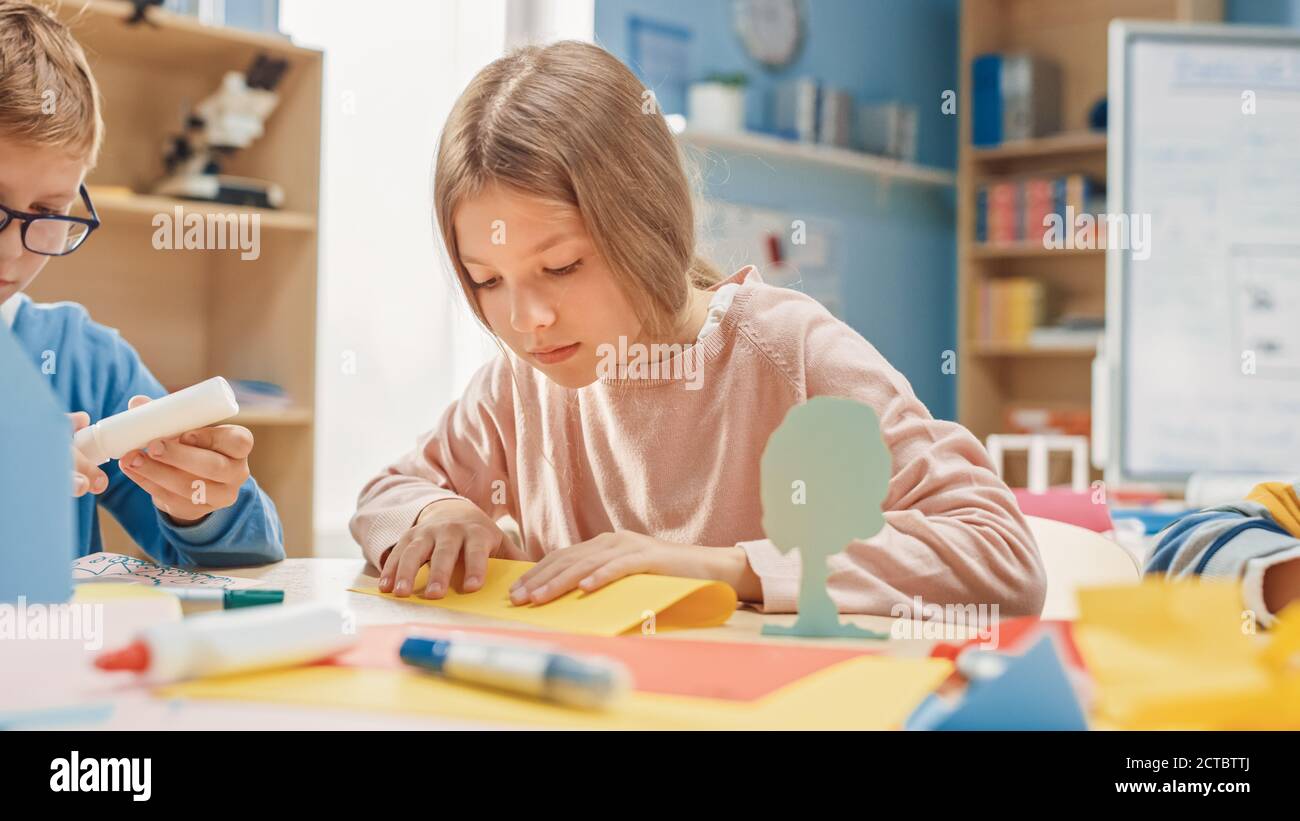Grundschule Kunst Handwerk Klasse: Niedliche kleine Blonde Mädchen hat Spaß mit ihren Klassenkameraden auf dem Handwerk-Projekt, verwendet buntes Papier zu falten Stockfoto