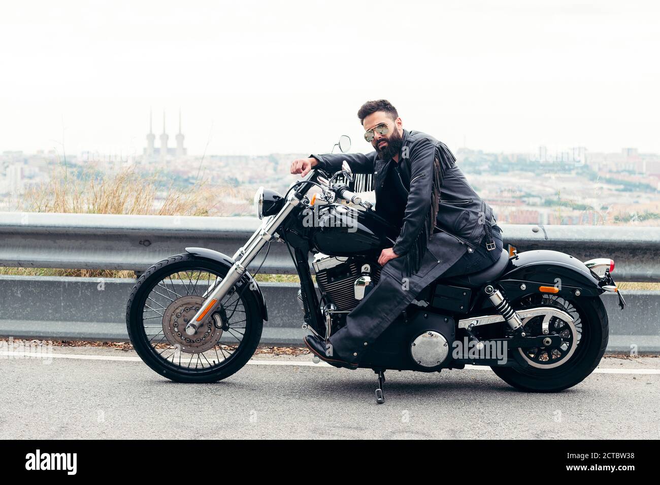 Mann in Leder und Sonnenbrille sitzend auf seinem Motorrad auf der Straße mit Panoramablick auf die Stadt, Konzept der Freiheit und Biker-Lifestyle, Stockfoto