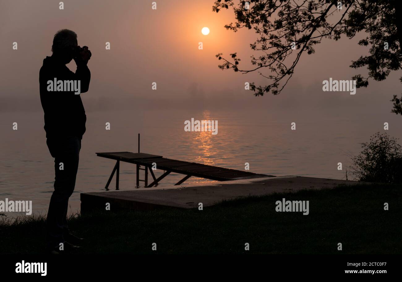 Silhouette Fotograf am hölzernen Steg bei Sonnenuntergang in der früh Morgen über dem Fluss maas in limburg in holland mit Die Bäume vernebeln und trüben Nebel Stockfoto