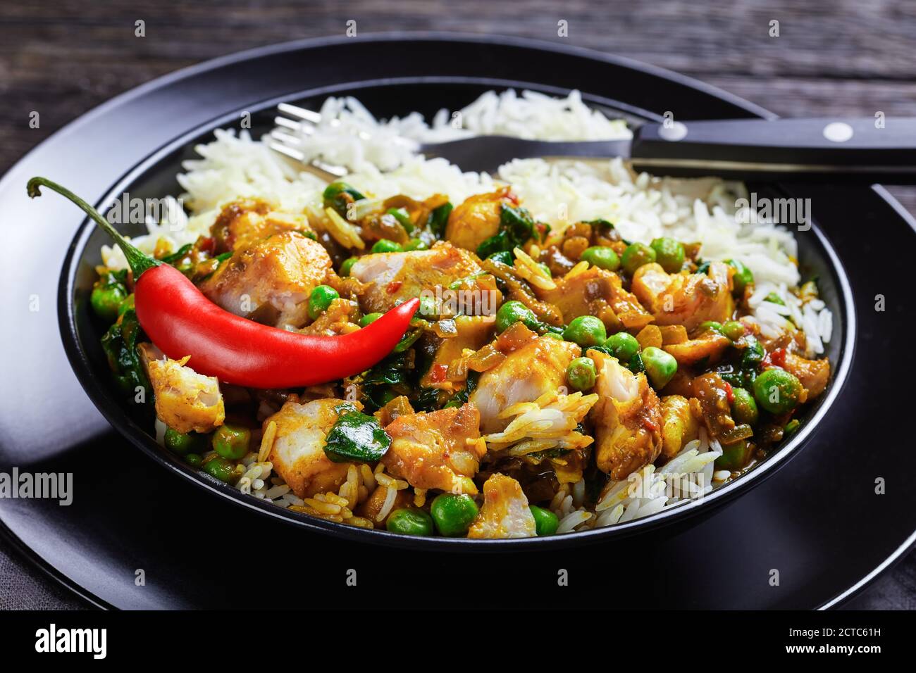 Fischcurry mit grünen Erbsen und Spinat serviert mit Reis in einer schwarzen Schüssel auf einem Holztisch, Landschaftsansicht von oben, Nahaufnahme Stockfoto