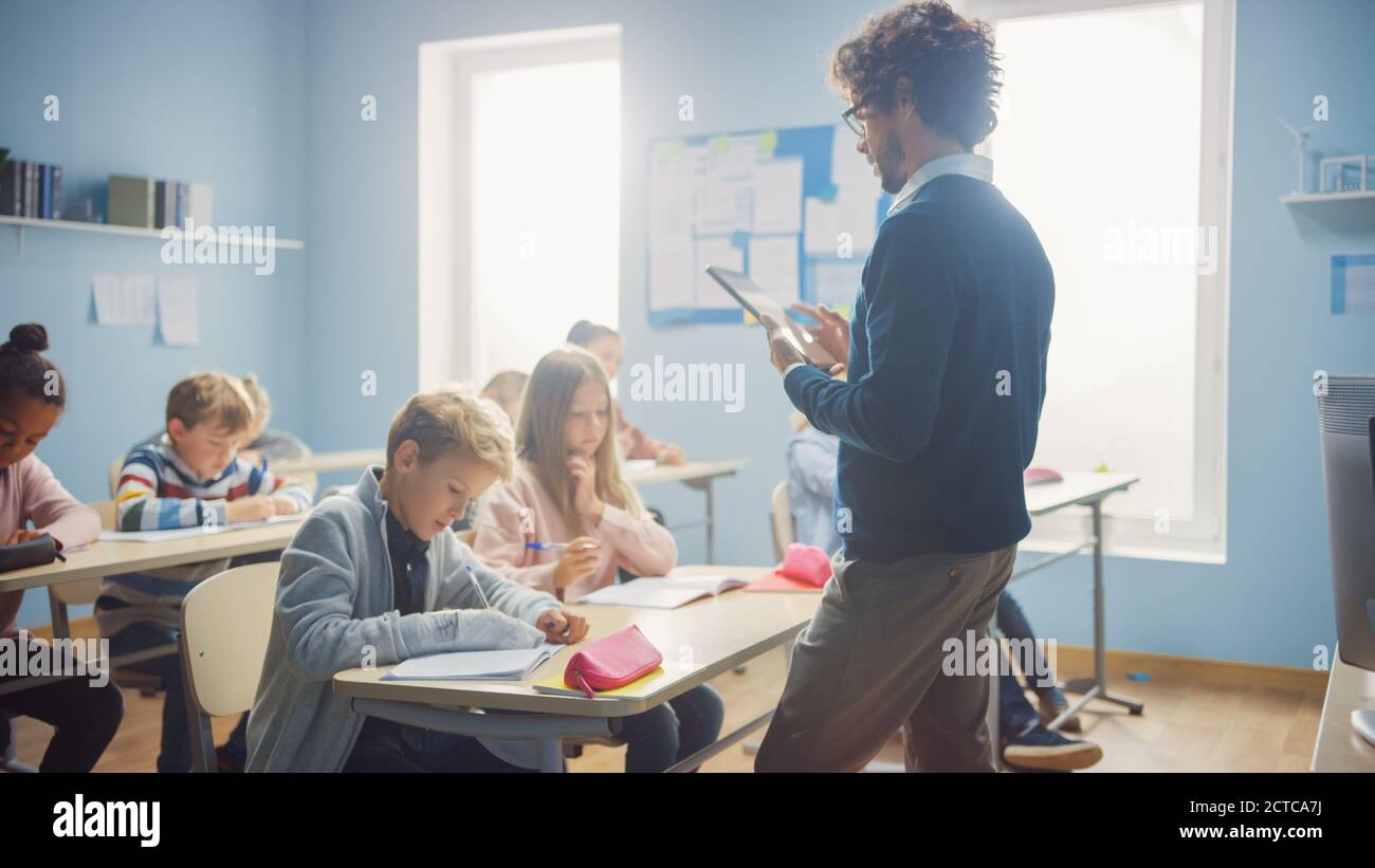 Der Lehrer verwendet einen digitalen Tablet-Computer, während er eine Lektion zu einem Klassenzimmer voller verschiedener Kinder erklärt, in der Grundschule Gruppe intelligenter multiethnischer Stockfoto
