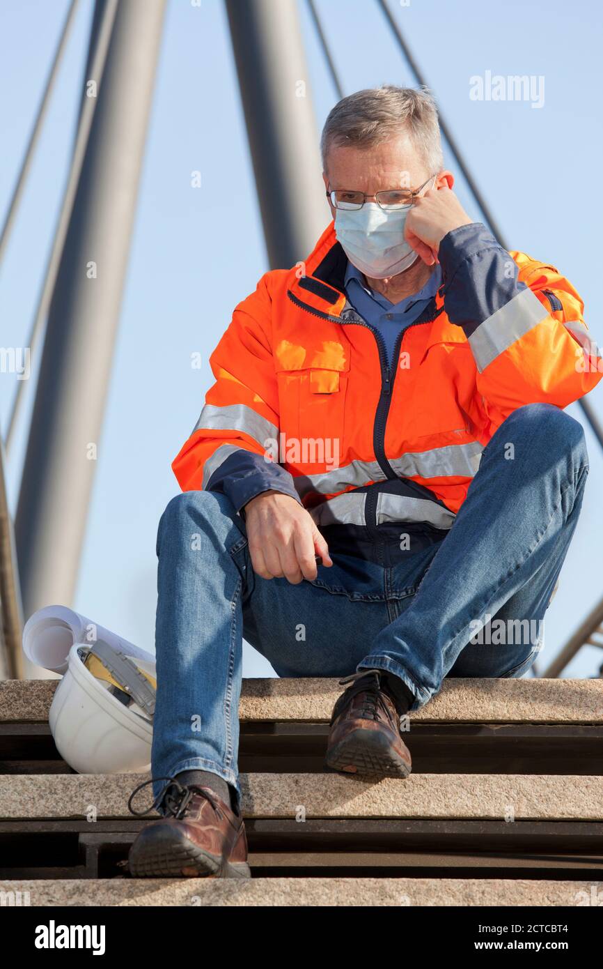 SAD Ingenieur mit Gesichtsmaske und schützender Arbeitskleidung sitzt in Vor einem industriellen Hintergrund - Fokus auf das Gesicht Stockfoto