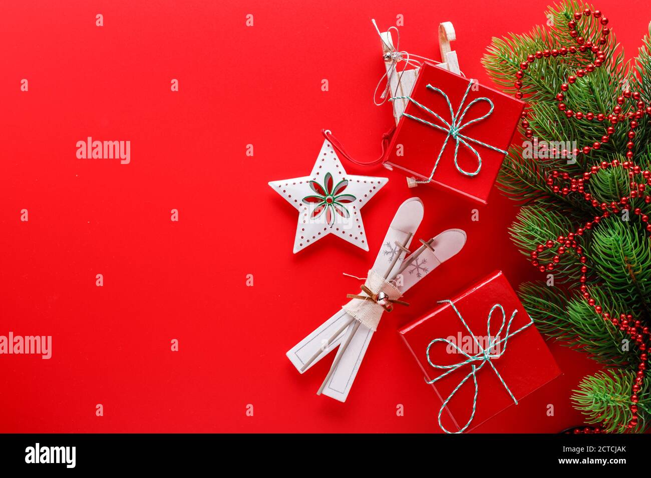 Draufsicht auf rotem Hintergrund mit Christbaumzweig, roten Geschenkschachteln, Sternornamenten und kleinen Holzskiern und Schlitten für die Weihnachtsfeiertage Stockfoto