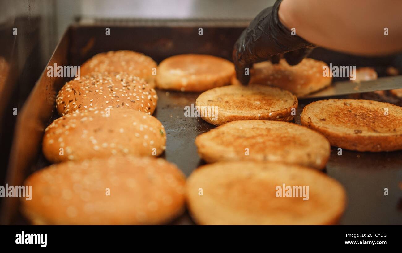 Schmackhafter Schuss eines Köchers, der Burger-Brötchen mit Sesamsamen auf einem Heißgas- oder Elektrogrill umdreht. Frische Gourmet Burger werden vorbereitet. Stockfoto