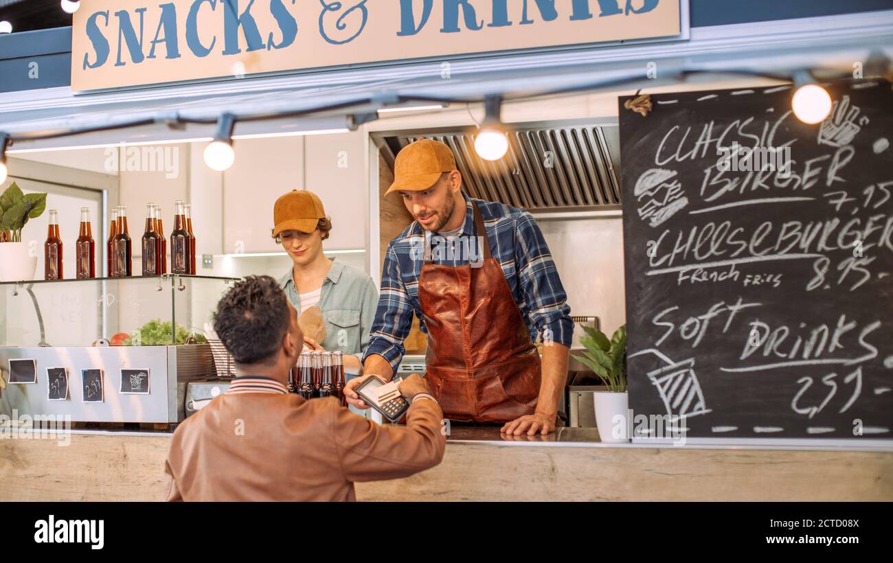 Junger indischer Mann in Lederjacke verwendet kontaktlose Bankkreditkarte, um für Nahrung zu zahlen. Street Food Truck Verkauf Burger Im Freien. Stockfoto