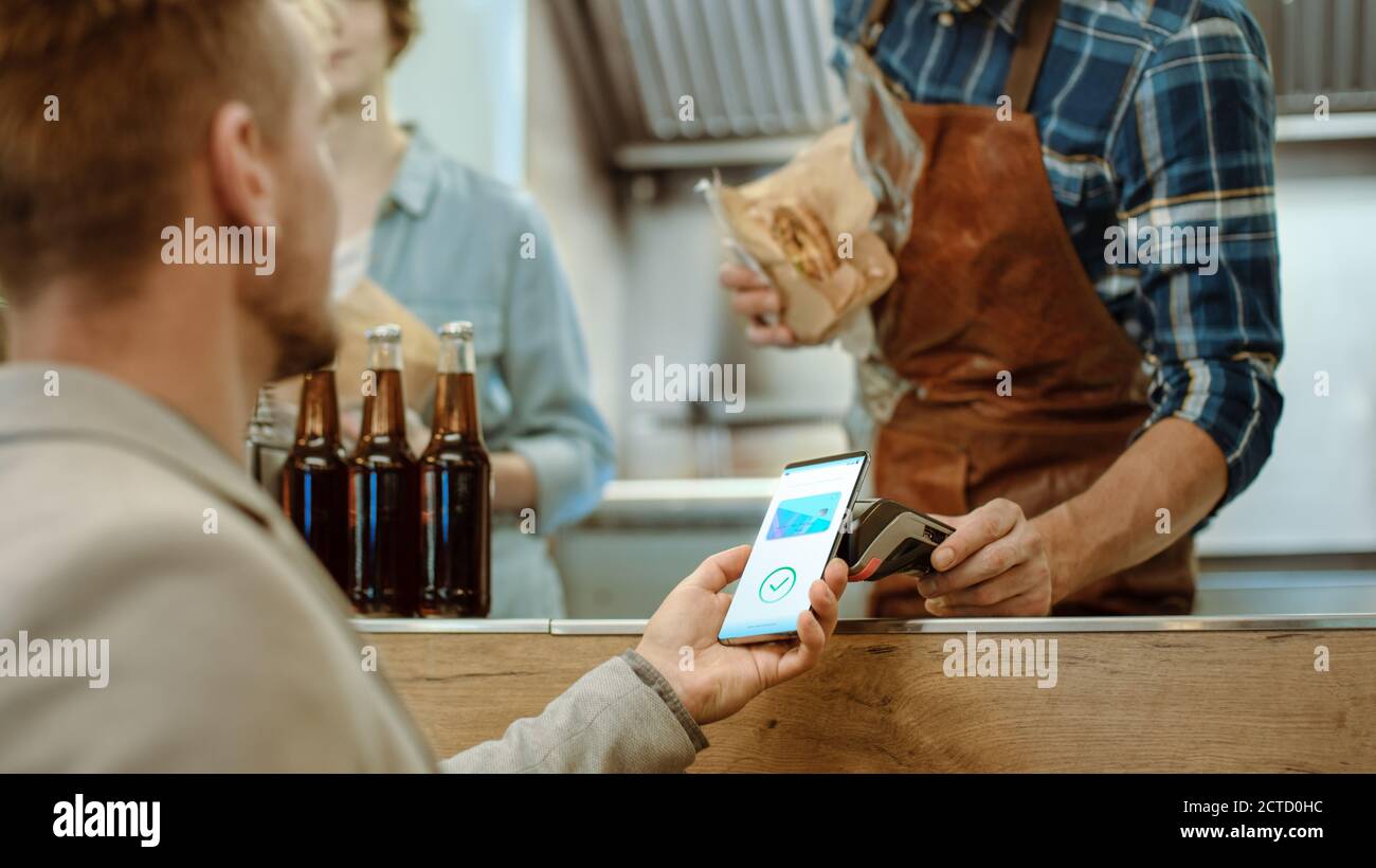 Food Truck Mitarbeiter gibt einen frisch gebackenen Burger an einen glücklichen jungen Mann. Mann in einem Casual Anzug verwendet sein Smartphone für NFC Mobile Payment Stockfoto