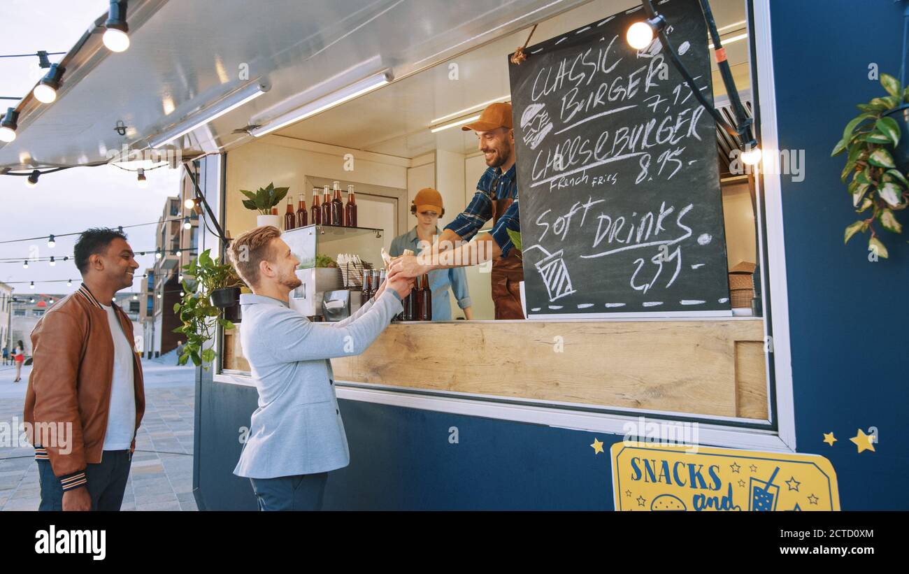 Food Truck Mitarbeiter gibt einem glücklichen jungen Mann in einem Anzug einen frisch zubereiteten Gourmet Burger. Street Food Truck Verkauf Burger in einem modernen Hip Stockfoto