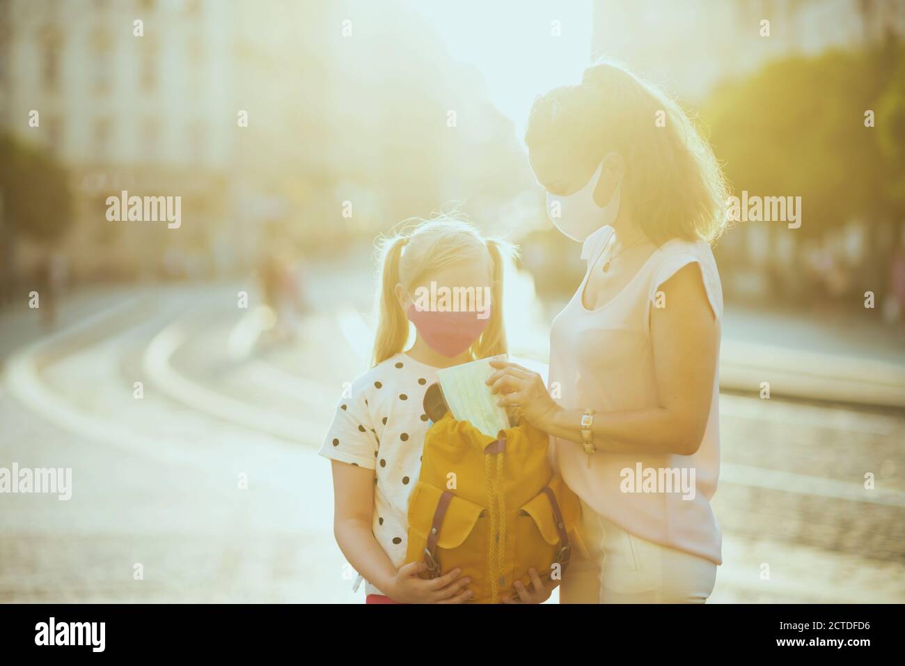 Leben während der Coronavirus-Pandemie. Elegante Mutter und Tochter mit Masken und gelben Rucksack Vorbereitung für die Schule und setzen zusätzliche medizinische Maske Stockfoto
