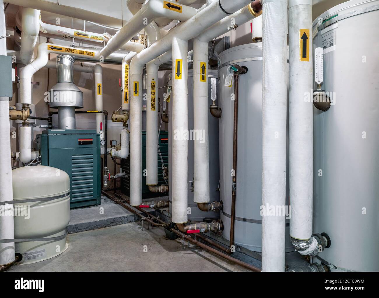 Kesselraum von Wohn-, Schichten- oder Mehrebenengebäude. 3 Warmwasserboiler sichtbar. Viele isolierte Rohre mit gelben Pfeilen in Richtung Wasser. Stockfoto
