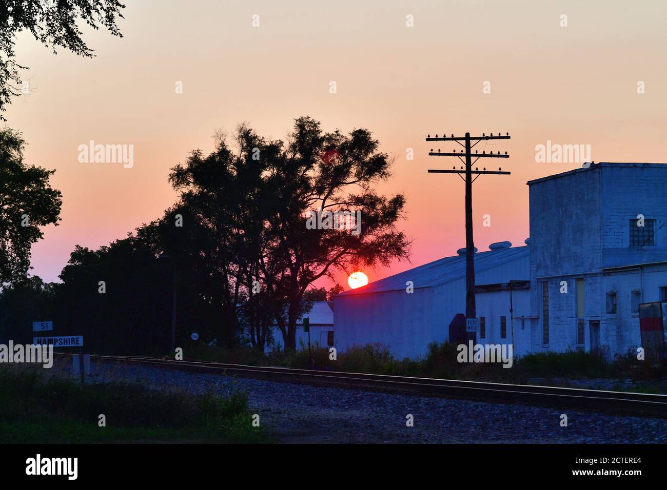 Hampshire, Illinois, USA. Der Sonnenuntergang hinter einigen Gebäuden und einer Eisenbahnstrecke entfacht ein farbenfrohes Ende eines Sommertages. Stockfoto