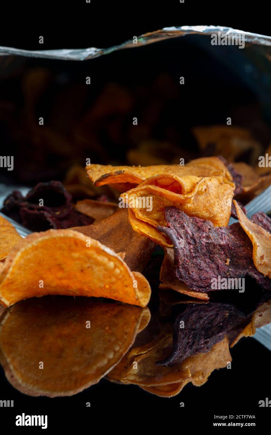 Beutel mit Gemüsechips auf schwarzem Hintergrund reflektiert. Geschmack von Süßkartoffeln, Rote Beete und Pastinak. Veganes Lebensmittelkonzept Stockfoto