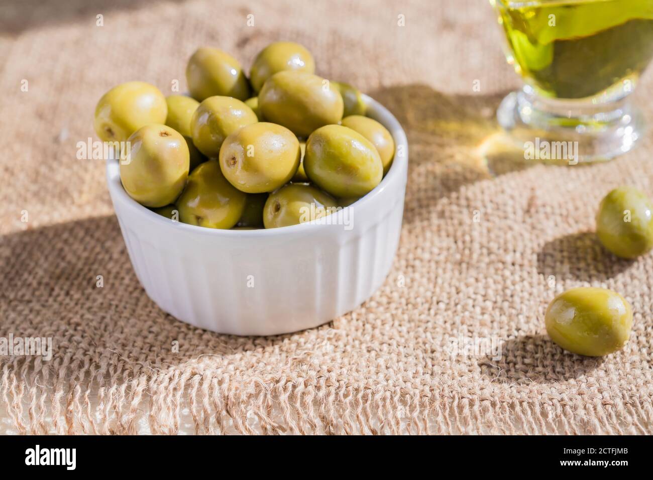 Frische marinierte grüne Oliven in weißer Keramikschale und Sauceboat Von hochwertigem nativem Olivenöl auf Sackleinen Stoff Hintergrund Stockfoto