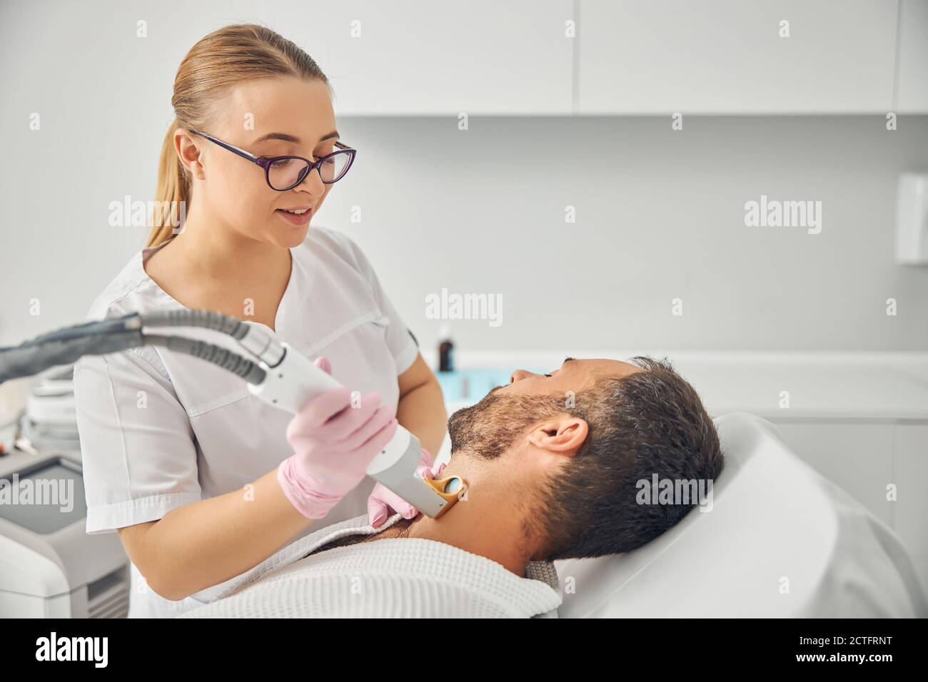 Charmante weibliche Ästhetikerin in sterilen Handschuhen entfernen unerwünschte Haare aus Männlicher Hals mit Lasergerät Stockfoto