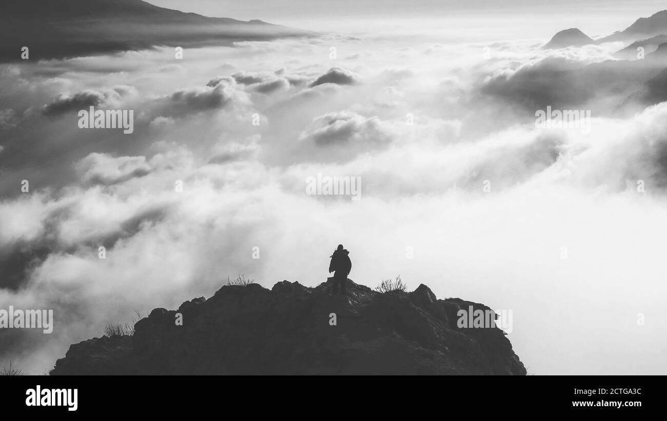 Eine Graustufenaufnahme eines einsamen Mannes, der auf dem steht Rand eines Berges mit Blick auf die Wolken bei Tageslicht Stockfoto