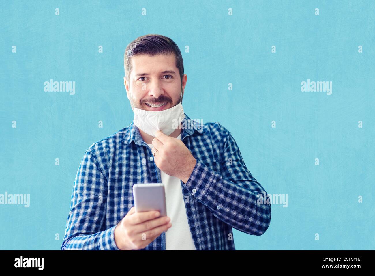 Selbstbewusster reifer Mann mit großem Lächeln und offenem Schutzgesicht Maske mit Smartphone gegen die Wand Stockfoto