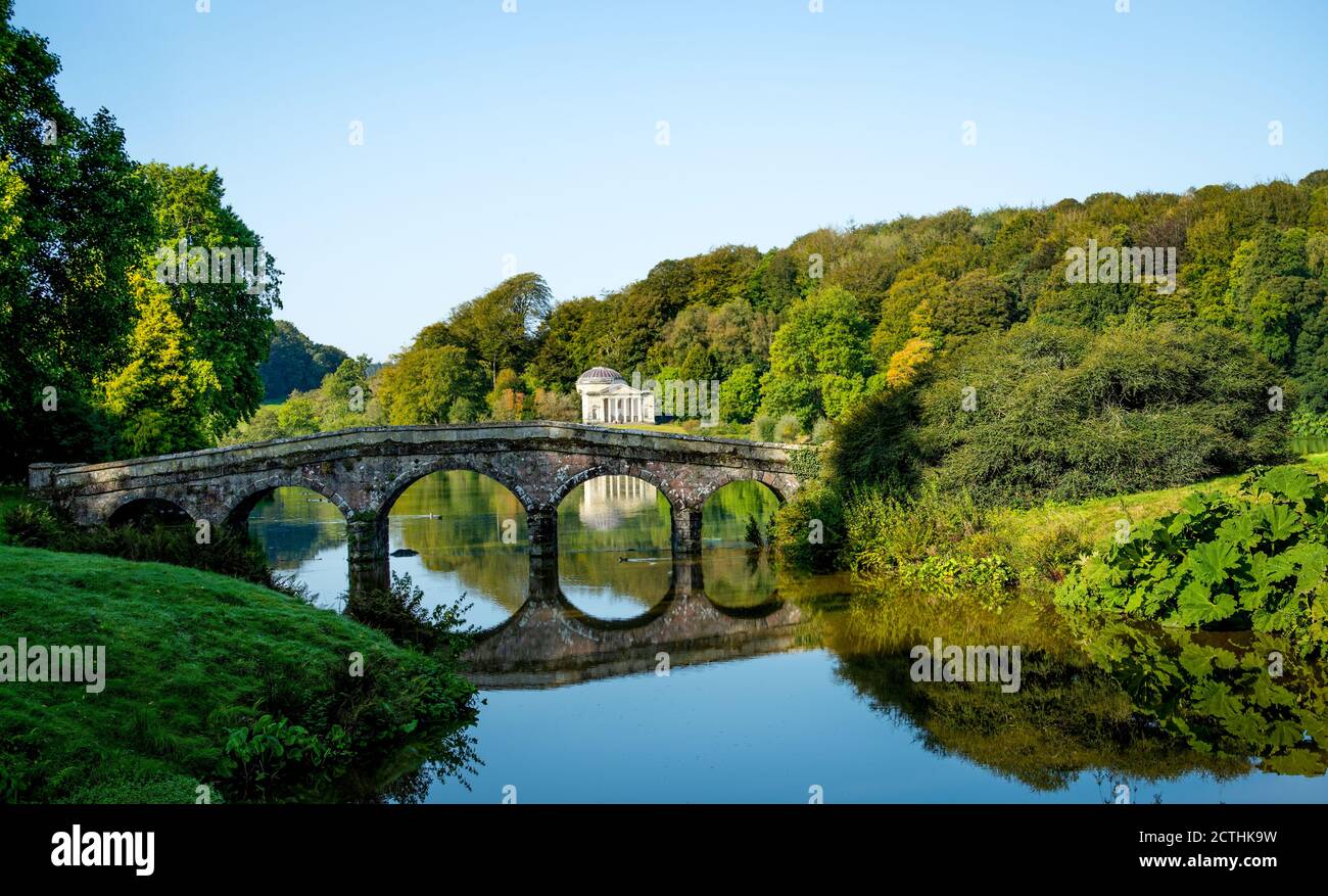 Die Palladio-Brücke, eine Steinbrücke mit fünf Bögen über das stille Wasser des Sees mit Bäumen, die beginnen, ihre Herbstblätter in den Stourhead-Gärten zu zeigen. Stockfoto