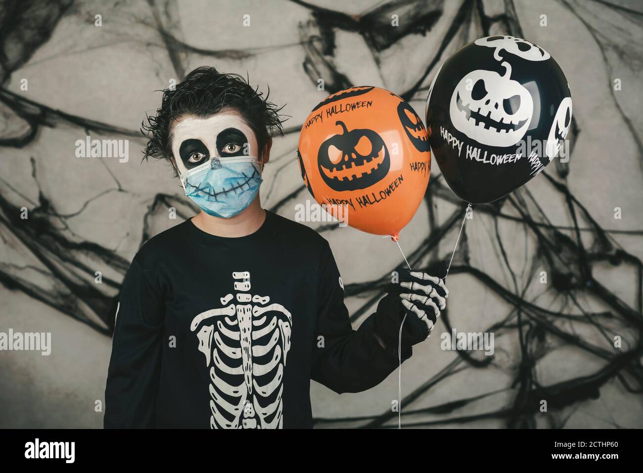 Happy Halloween. Kind mit medizinischer Maske in einem Skelett Kostüm mit halloween Ballons auf grauem Hintergrund Stockfoto