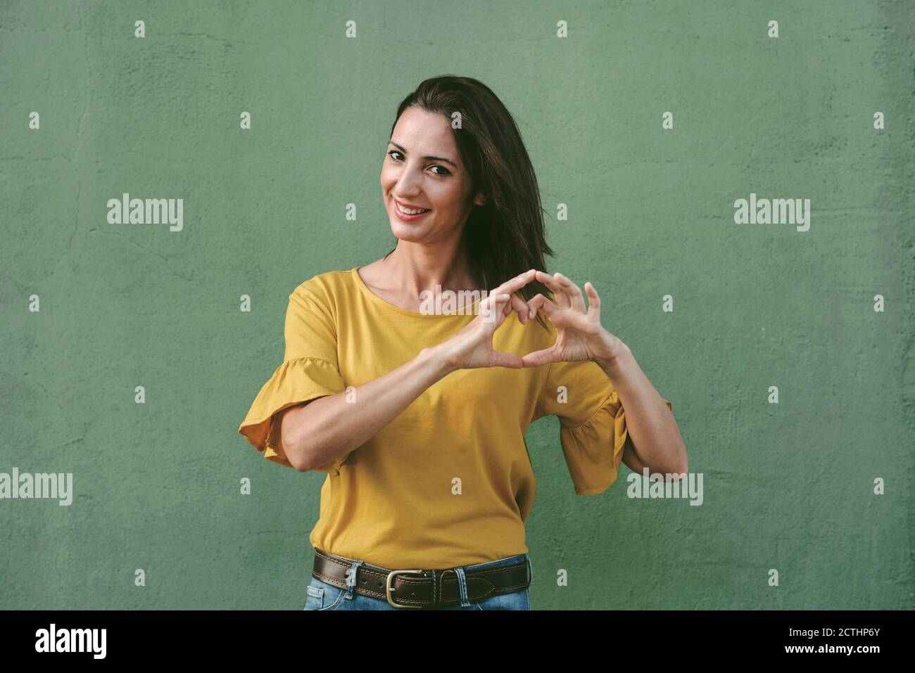 Lächelnde junge Frau zeigt Herz mit Händen über grünem Hintergrund Stockfoto