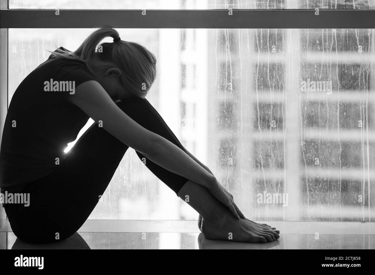 Traurigkeit und Verzweiflung - deprimierte junge Frau, die allein zu Hause am Fenster mit Blick auf die Stadt sitzt. Schwarz und Weiß. Stockfoto