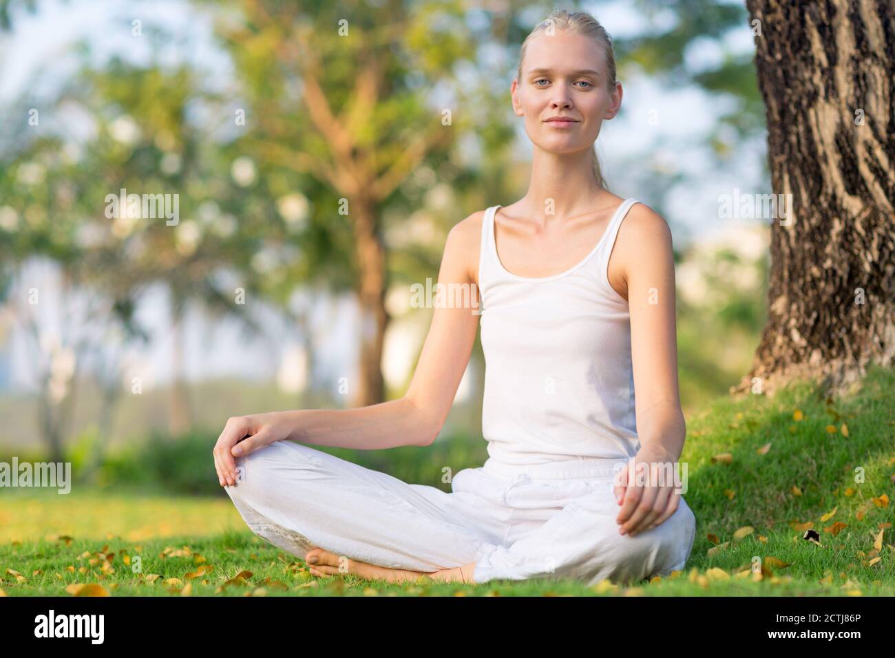 Schöne, ruhige Frau, die in einem ruhigen grünen Park sitzt, während sie zur Kamera lächelt. Wohlbefinden und Selbstversorgung. Stockfoto