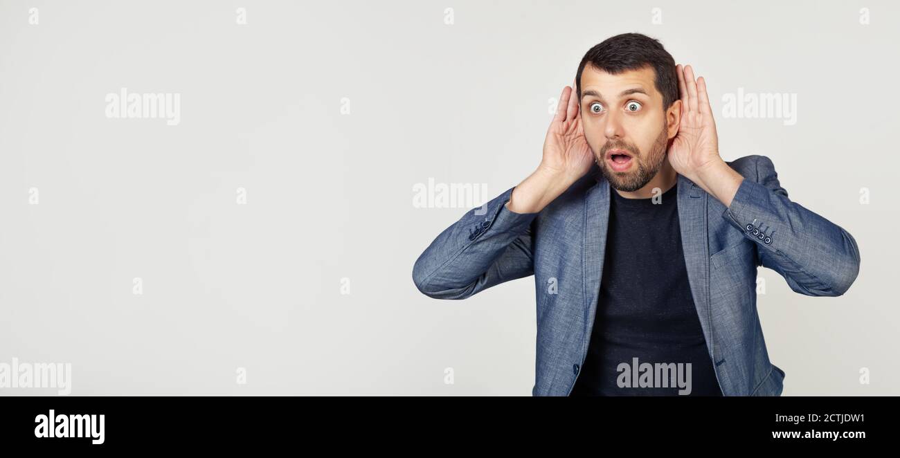 Junger Geschäftsmann Mann mit Bart versucht, beide Hände auf seinem Ohr zu hören, neugierig auf Klatsch. Hörprobleme, taub. Porträt eines Mannes auf grauem Hintergrund. Stockfoto