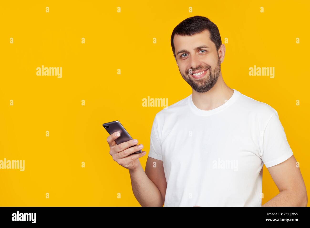 Der junge Mann mit einem Bart in einem weißen T-Shirt benutzt ein Smartphone mit einem glücklichen Gesicht, das steht und lächelt und ein selbstbewusstes Lächeln zeigt, das Zähne zeigt. Steht auf isoliertem gelben Hintergrund. Stockfoto