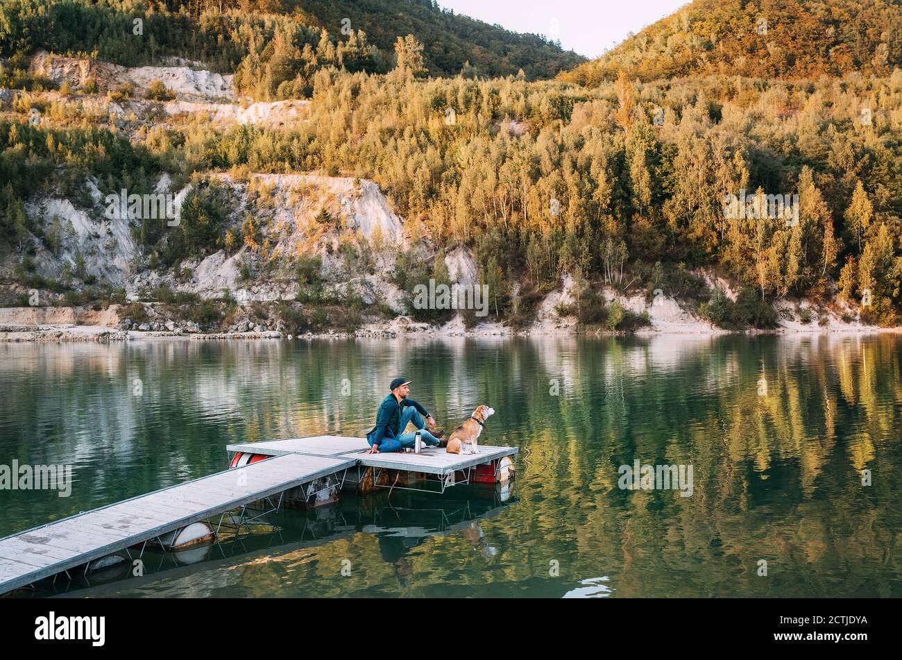 Mann als Hundebesitzer und sein Freund beagle Hund Sitzen auf dem hölzernen Pier auf dem Bergsee Und genießen Sie die Landschaft während ihrer Wanderung in Th Stockfoto