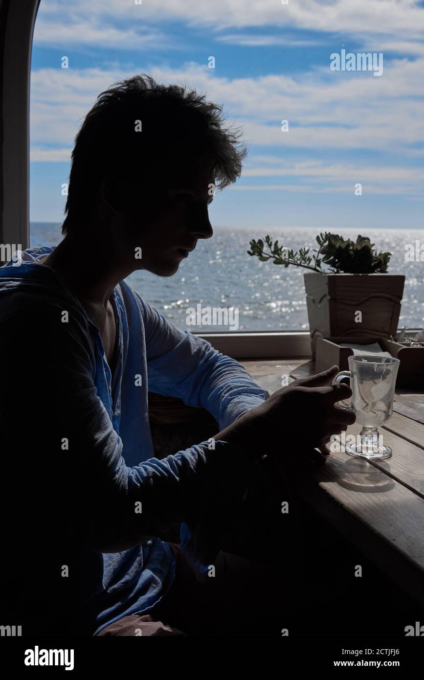 Silhouette eines jungen Mannes, der einen Cappuccino vor dem Hintergrund eines Fensters mit Meerblick beendet. Stockfoto