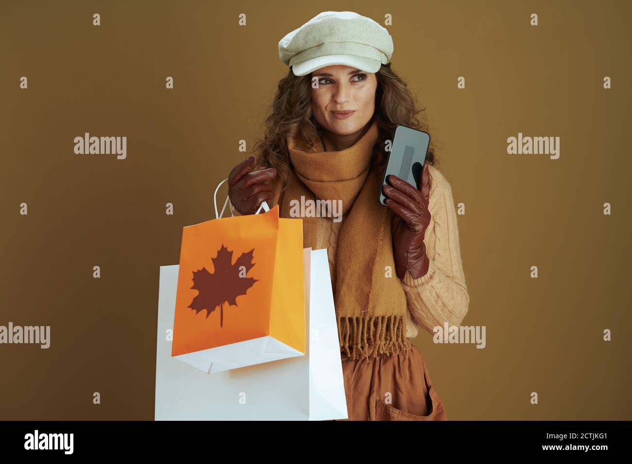 Hallo Herbst. Nachdenkliche trendige Hausfrau mittleren Alters in Pullover mit Smartphone, Lederhandschuhe und Papier Einkaufstasche vor braunem Hintergrund. Stockfoto