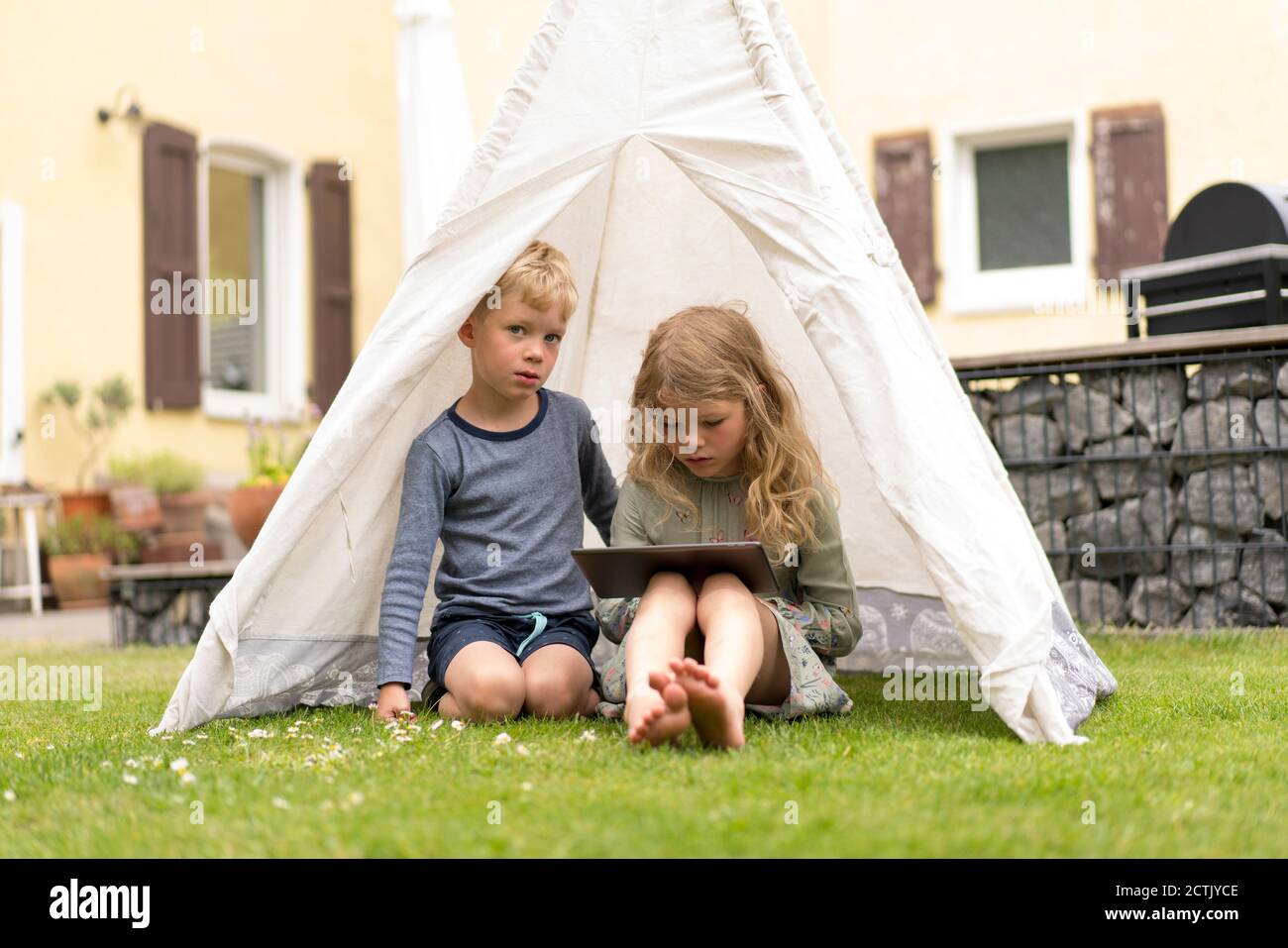 Nette Geschwister mit digitalen Tablet sitzen im Zelt auf Gras Auf Hinterhof gegen Haus Stockfoto