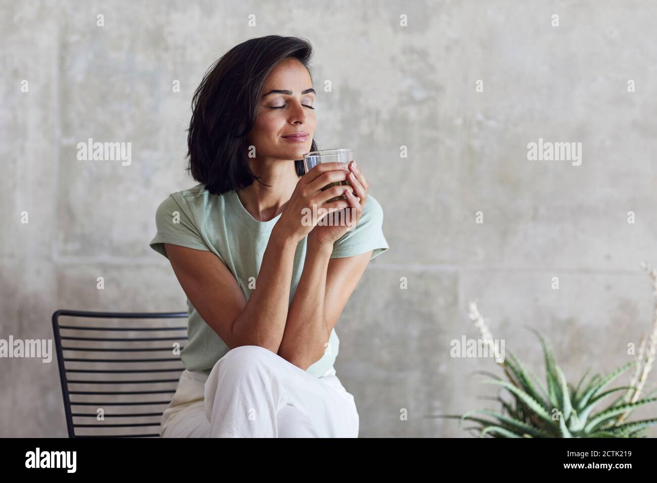 Geschäftsfrau mit geschlossenen Augen hält Smoothie, während sie auf einem Stuhl sitzt Gegen die Wand im Büro Stockfoto