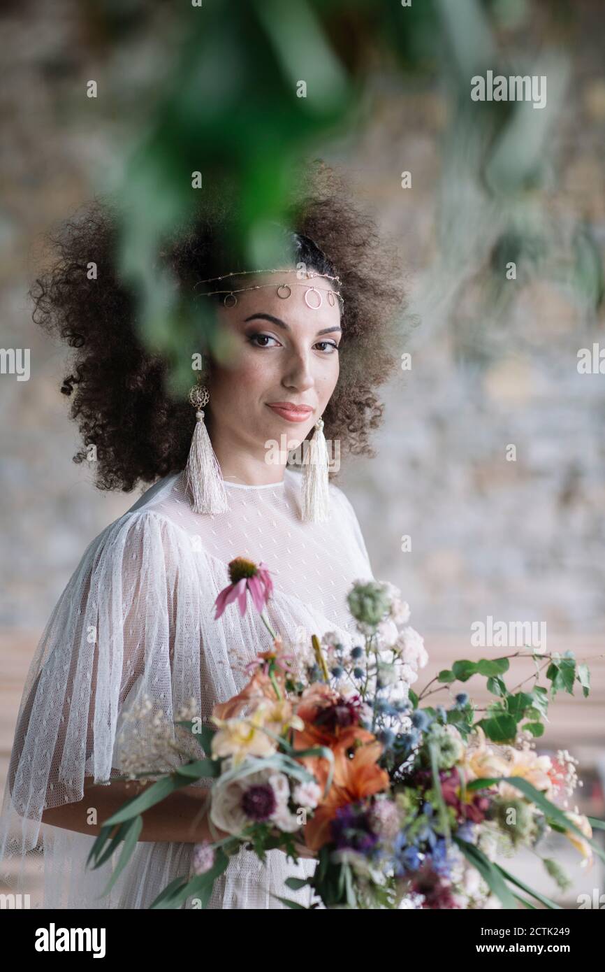 Junge Frau im Hochzeitskleid hält Strauß beim Stehen gegen Wand Stockfoto