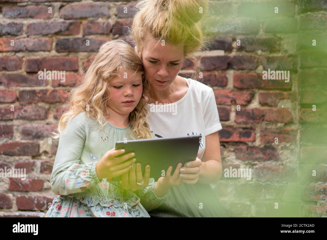 Frau und Tochter betrachten digitale Tablette, während sie gegen sitzen Backsteinmauer im Hinterhof Stockfoto