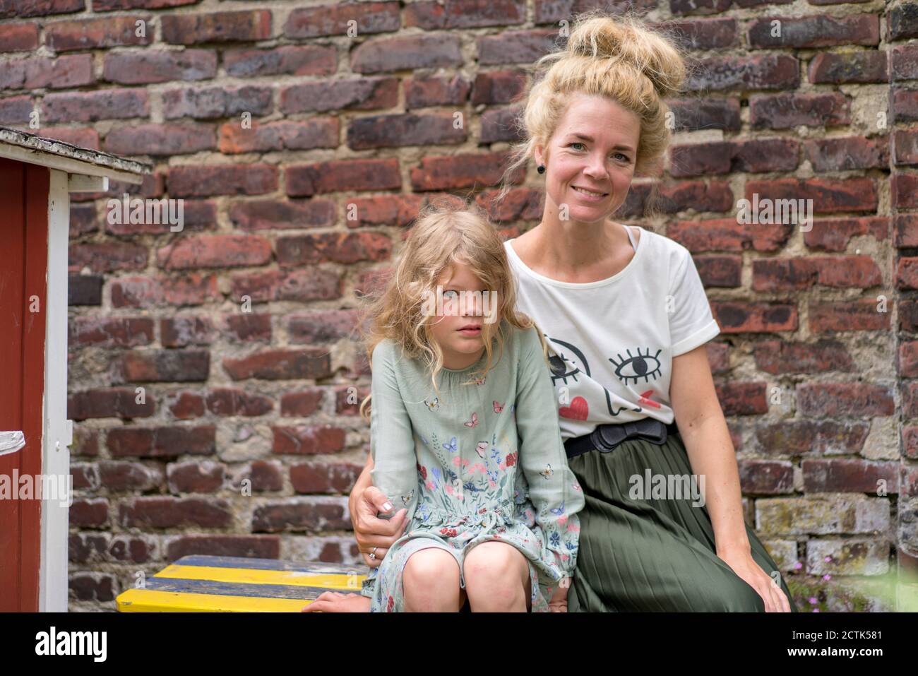 Lächelnde Frau mit Tochter mit blonden Haaren, die gegen Backstein sitzen Wand im Hinterhof Stockfoto