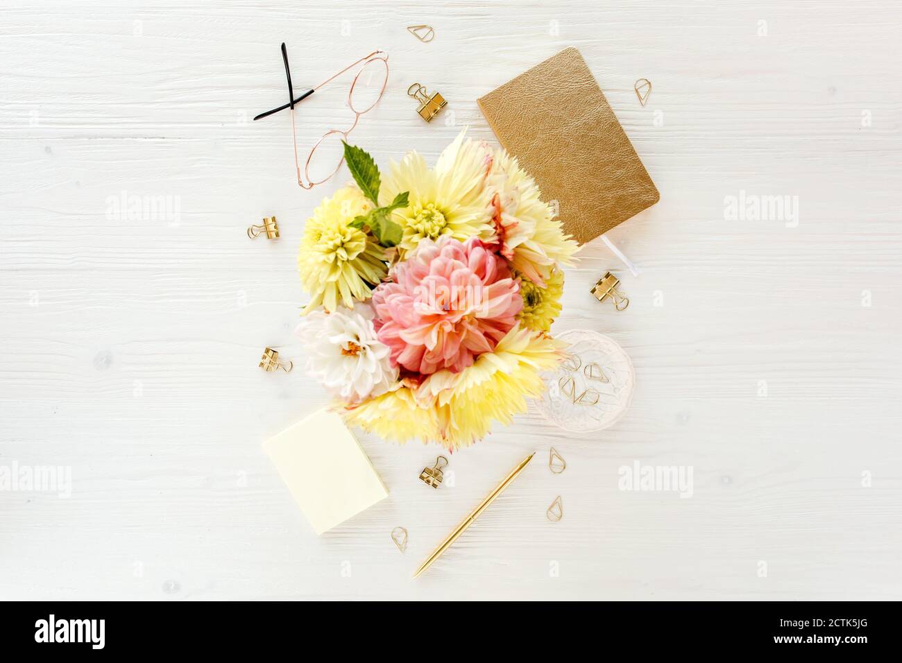 Flacher Schreibtisch für Damen. Weiblicher Arbeitsplatz mit gelben Dahlien und Rosenblüten, Accessoires, goldenem Tagebuch, weißem Holzhintergrund. Draufsicht. Stockfoto