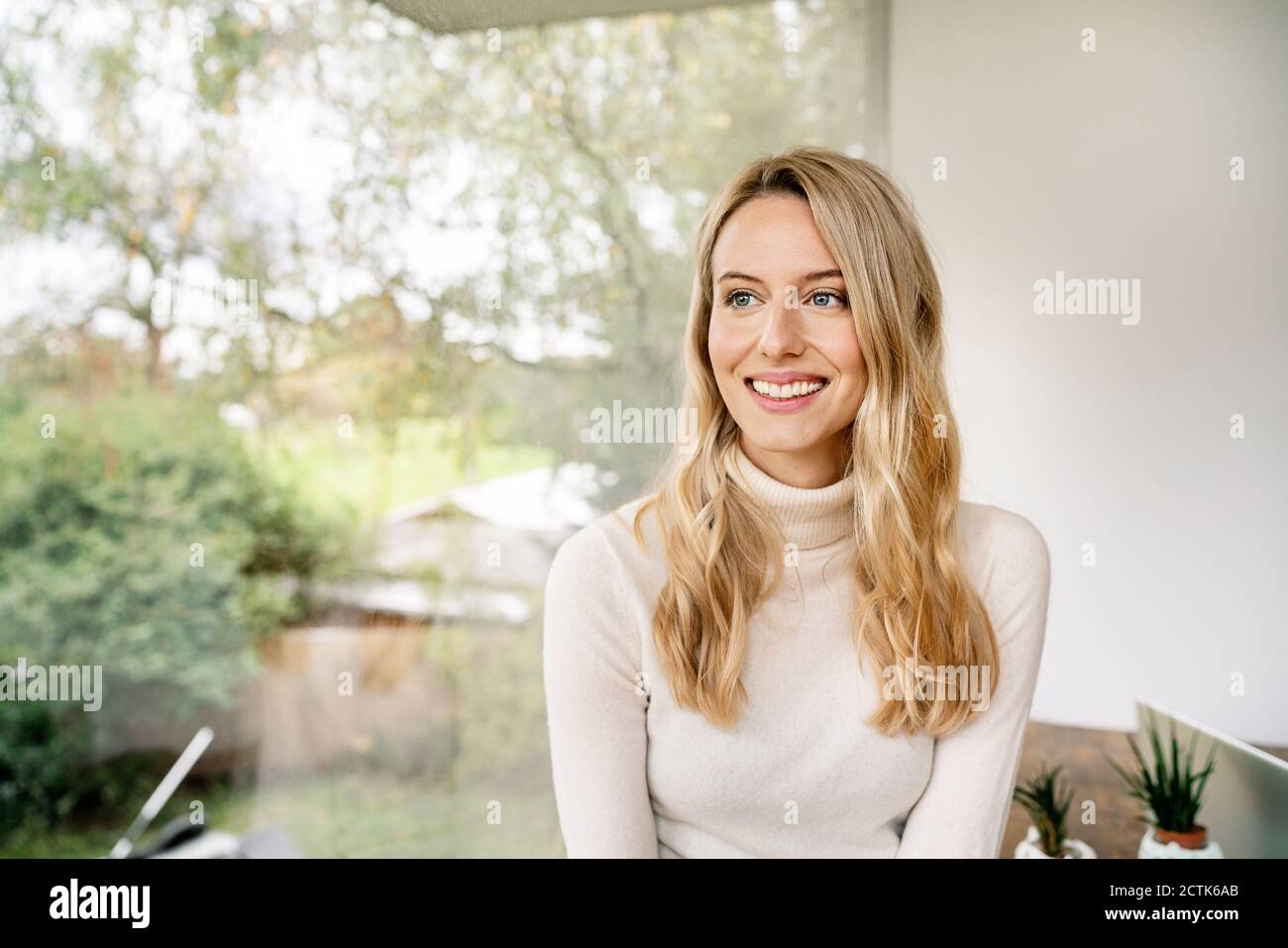 Lächelnde, nachdenkliche Geschäftsfrau mit blonden Haaren, die am Fenster sitzen Büro Stockfoto