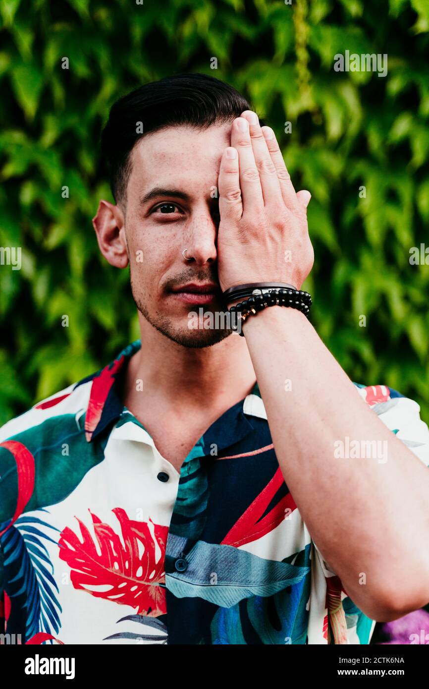 Mann, der das Gesicht mit einer Hand bedeckt, während er gegen Efeu steht Hecke Stockfoto