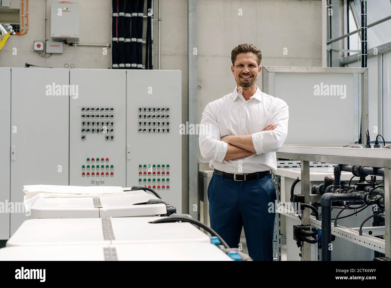 Lächelnder Geschäftsmann mit gekreuzten Armen, die im Gewächshaus von der Ausrüstung stehen Stockfoto