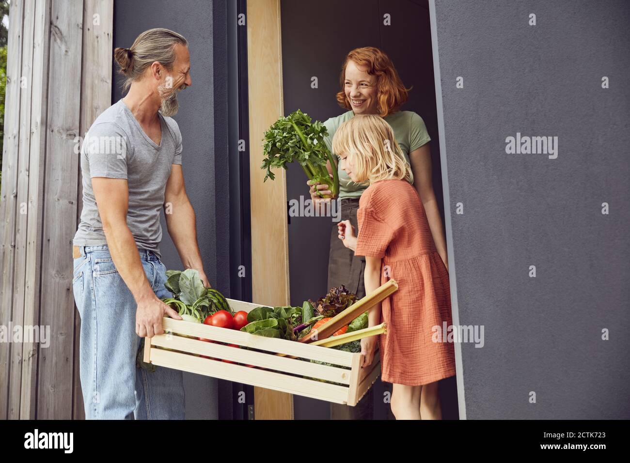 Lächelnde Frau hält Gemüse, während Vater und Tochter Kiste tragen Am Eingang Stockfoto
