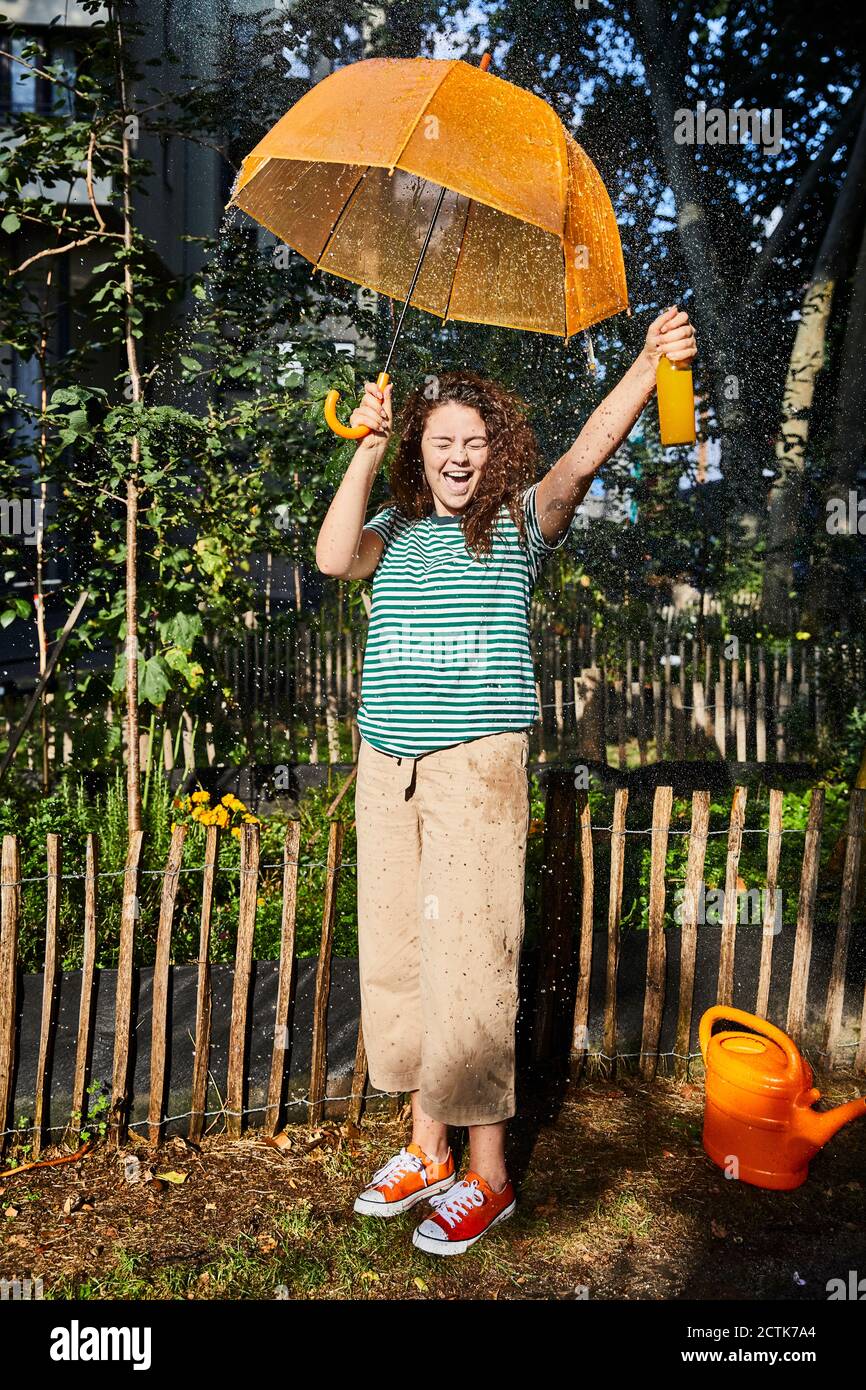 Fröhliche Frau mit Mund offen hält Flasche und Regenschirm während Stehen auf Hinterhof genießen sonnigen Tag Stockfoto