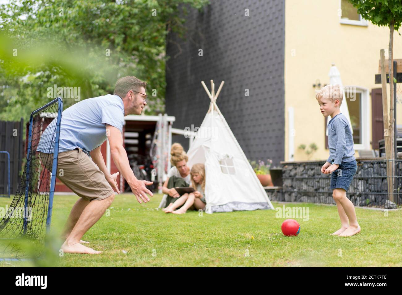 Mann und Sohn spielen Fußball, während Frau mit Tochter sitzt Im Zelt im Hinterhof Stockfoto