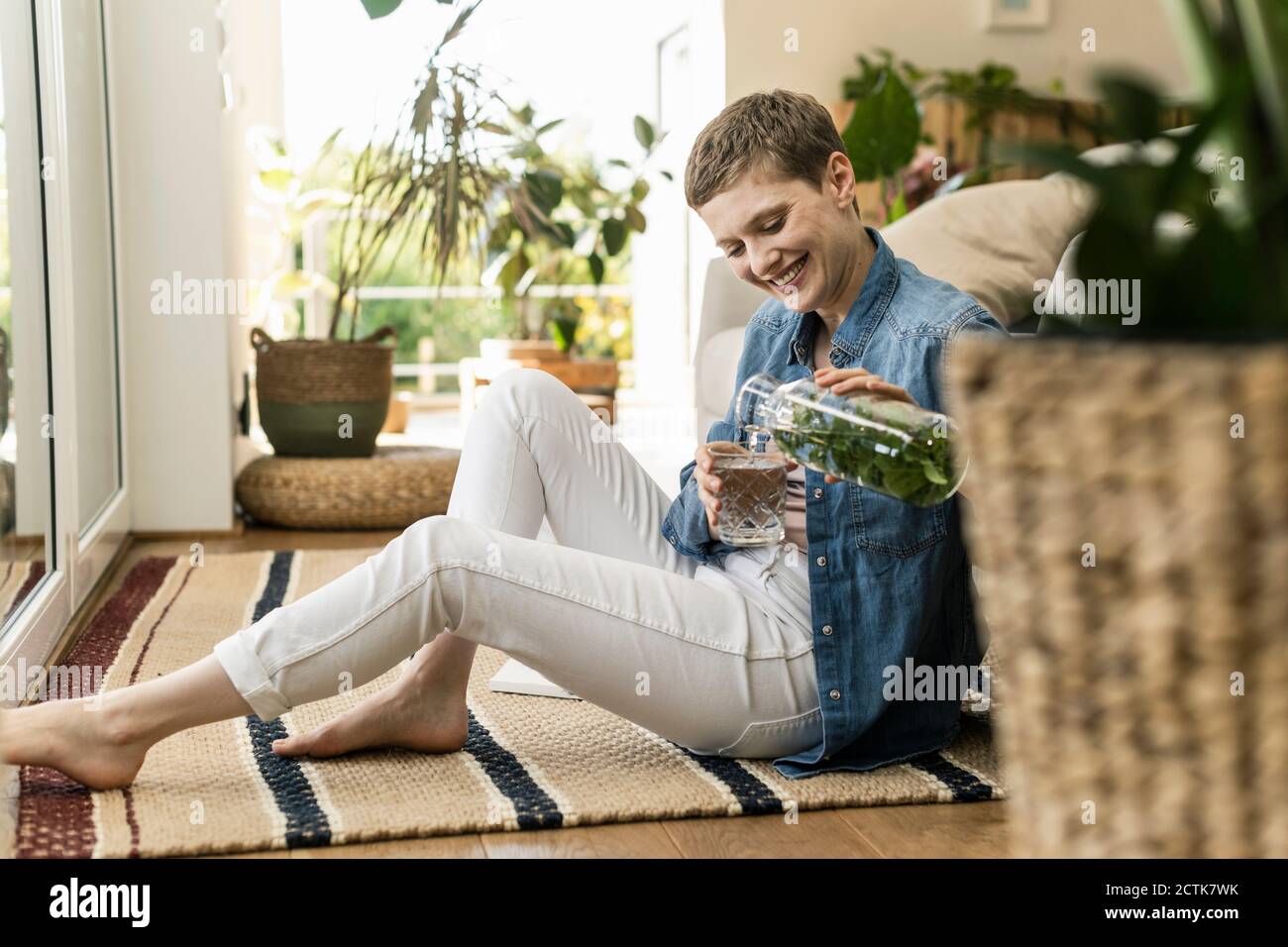 Lächelnde Frau mit kurzen Haaren Gießen trinken in Glas, während Zu Hause auf dem Teppich sitzen Stockfoto