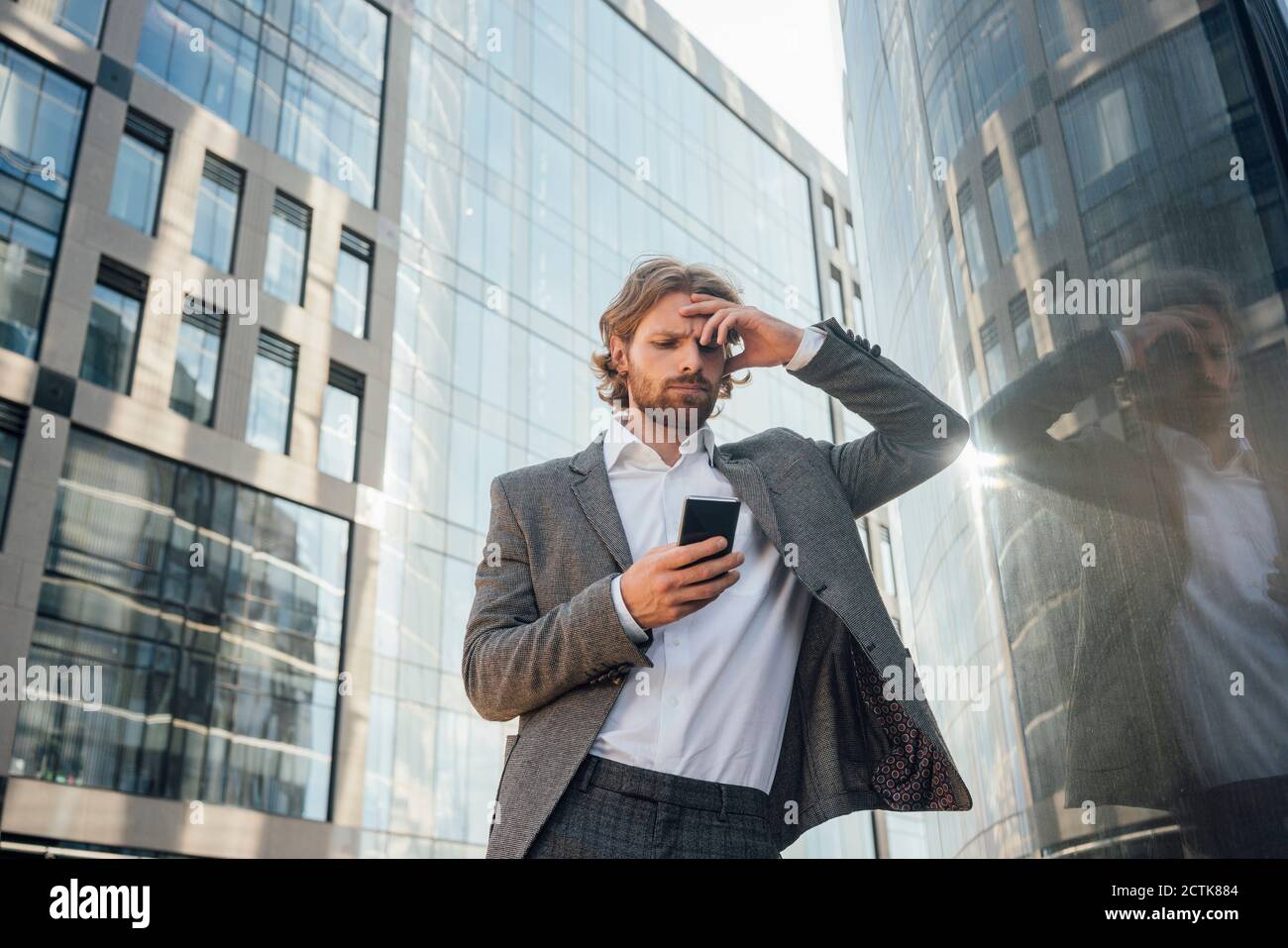 Besorgt männlichen Profi mit Kopf in der Hand mit Smartphone Während Sie sich an einem Bürogebäude in der Innenstadt lehnten Stockfoto