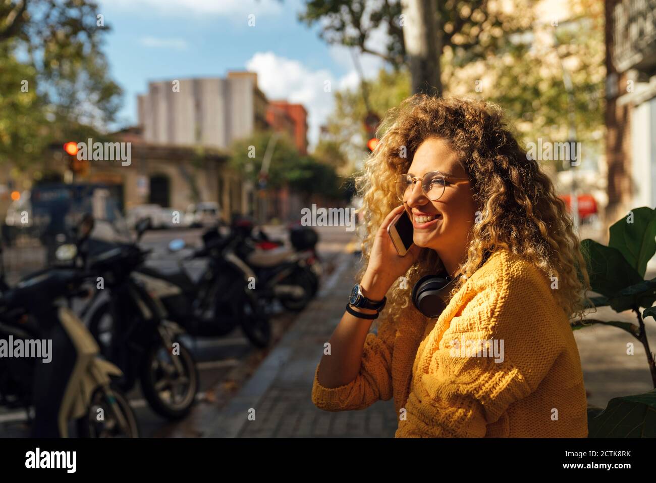 Glückliche junge Studentin mit lockigen blonden Haaren im Gespräch Smartphone in der Stadt Stockfoto