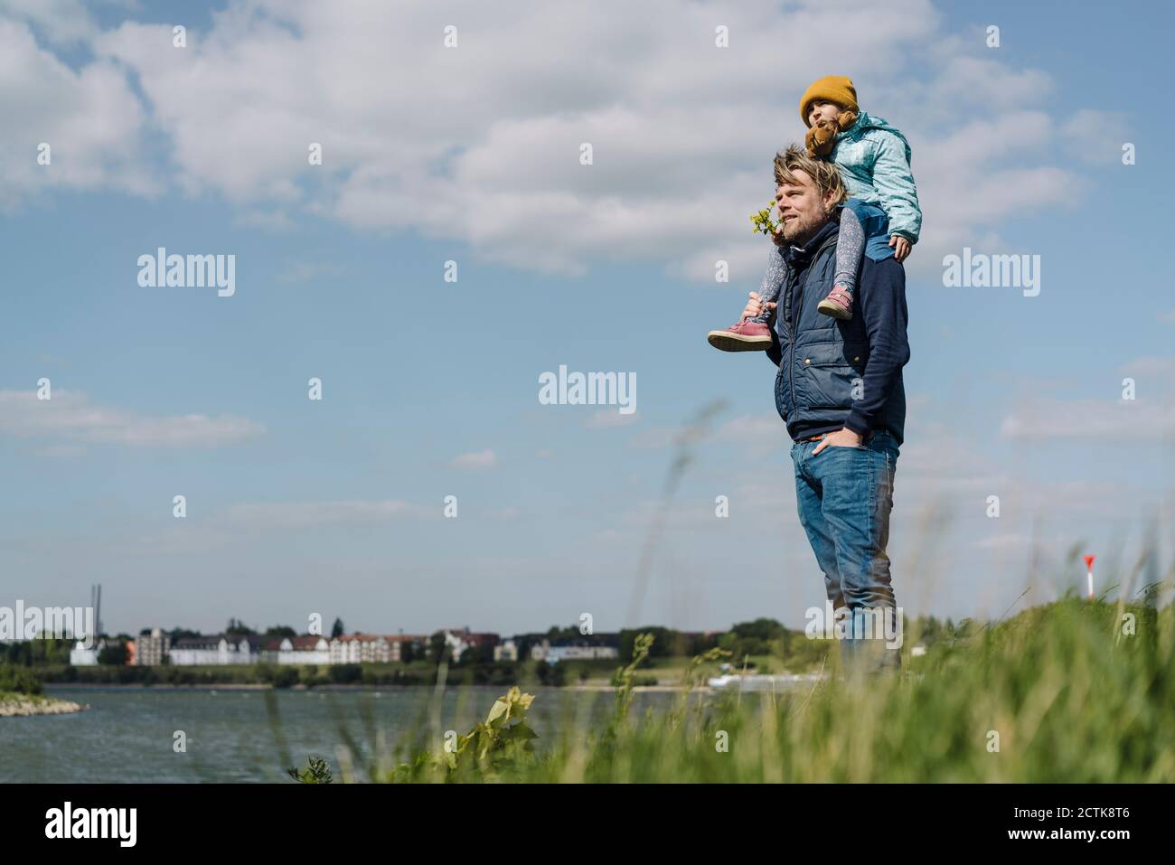 Vater steht mit Händen in den Taschen, während er die Tochter trägt Schulter in der Nähe des Rheins Stockfoto