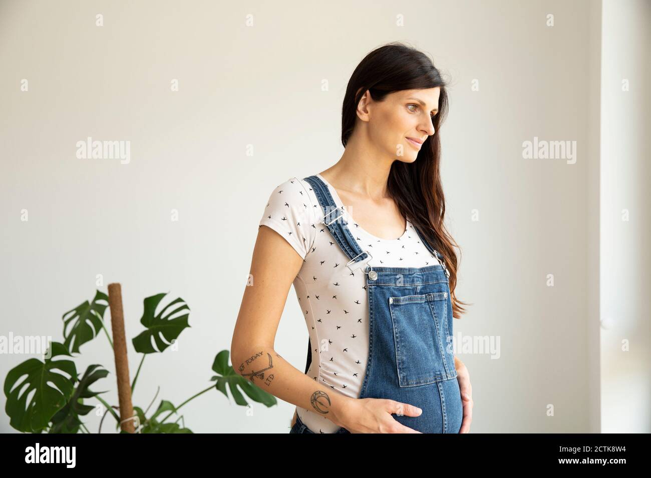 Nachdenkliche schwangere Frau mit Händen auf dem Bauch, die gegen die Wand stehen In neuem Zuhause Stockfoto