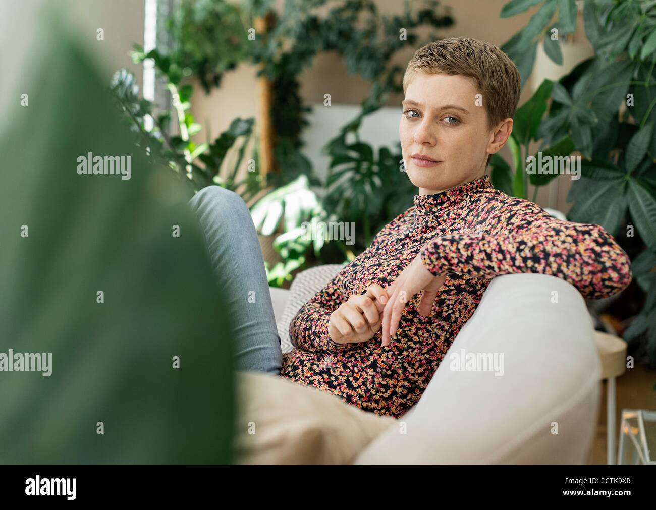 Selbstbewusst schöne Frau mit kurzen Haaren sitzen auf dem Sofa gegen Zimmerpflanzen zu Hause Stockfoto