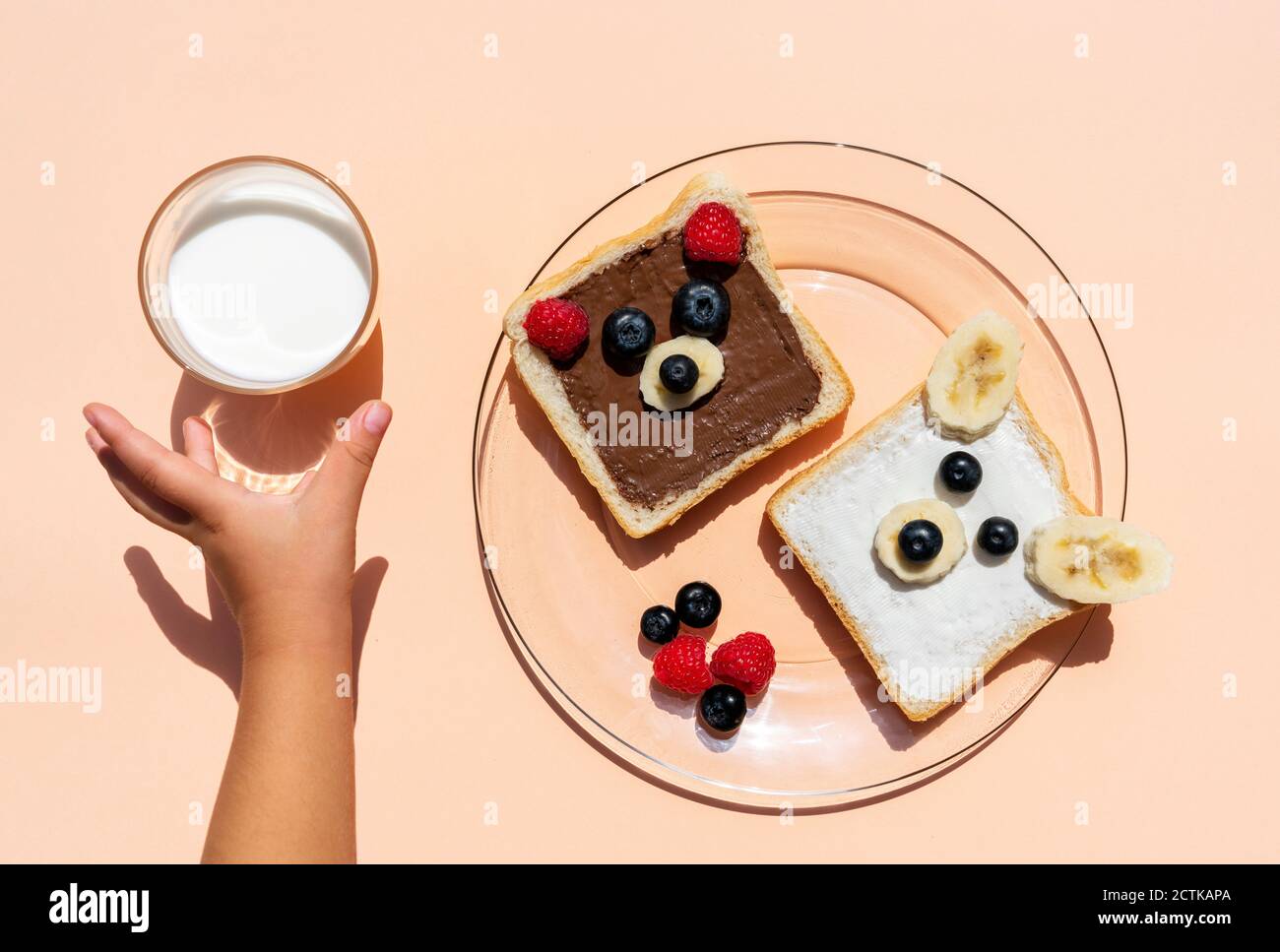 Studioaufnahme von Toasts mit Bärengesichtern aus Früchten Und die Hand des kleinen Mädchens, das nach einem Glas Milch greift Stockfoto