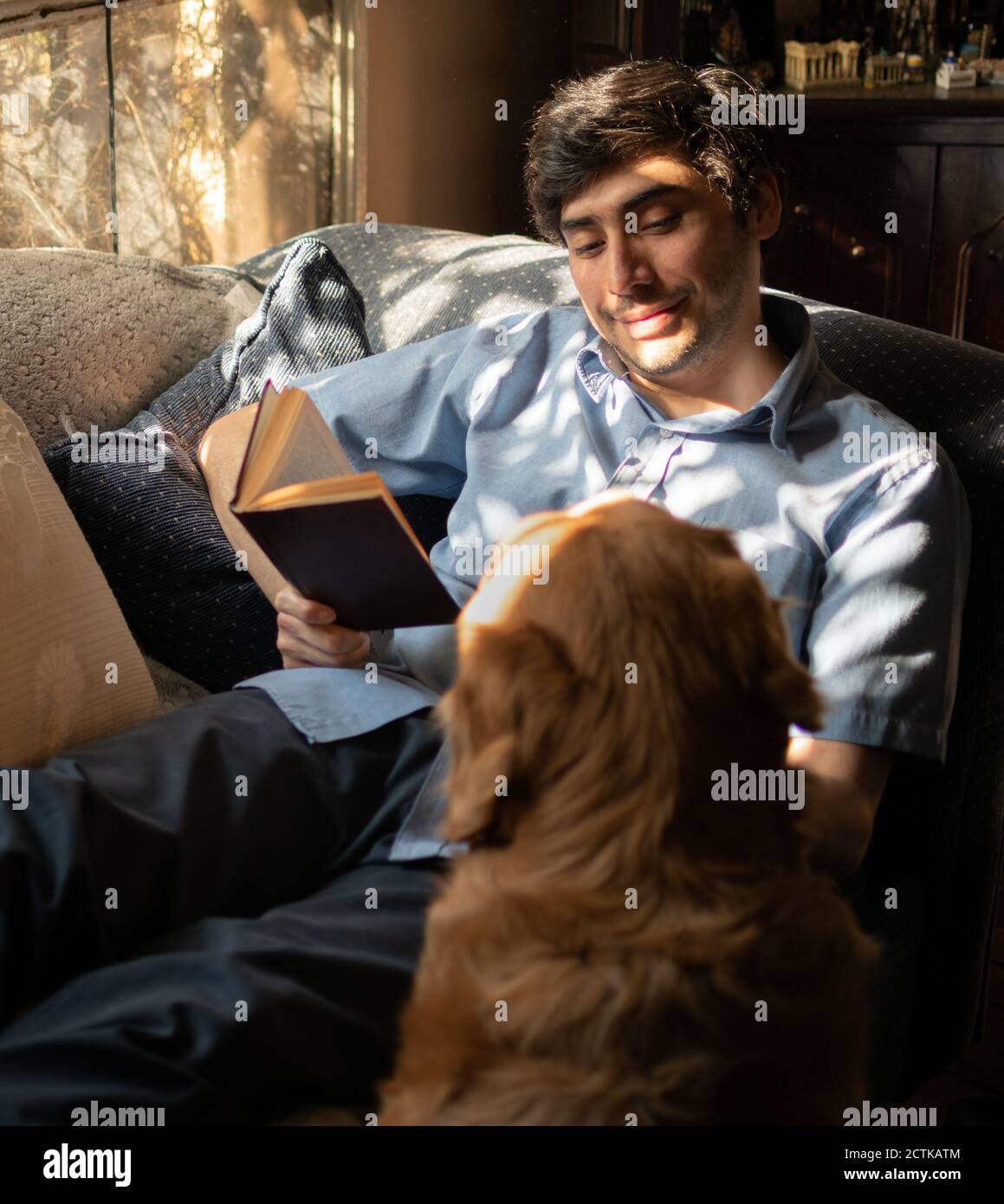 Ein entspannter Mann liest ein Buch und liegt auf einem Sofa neben einem Hund. Stockfoto